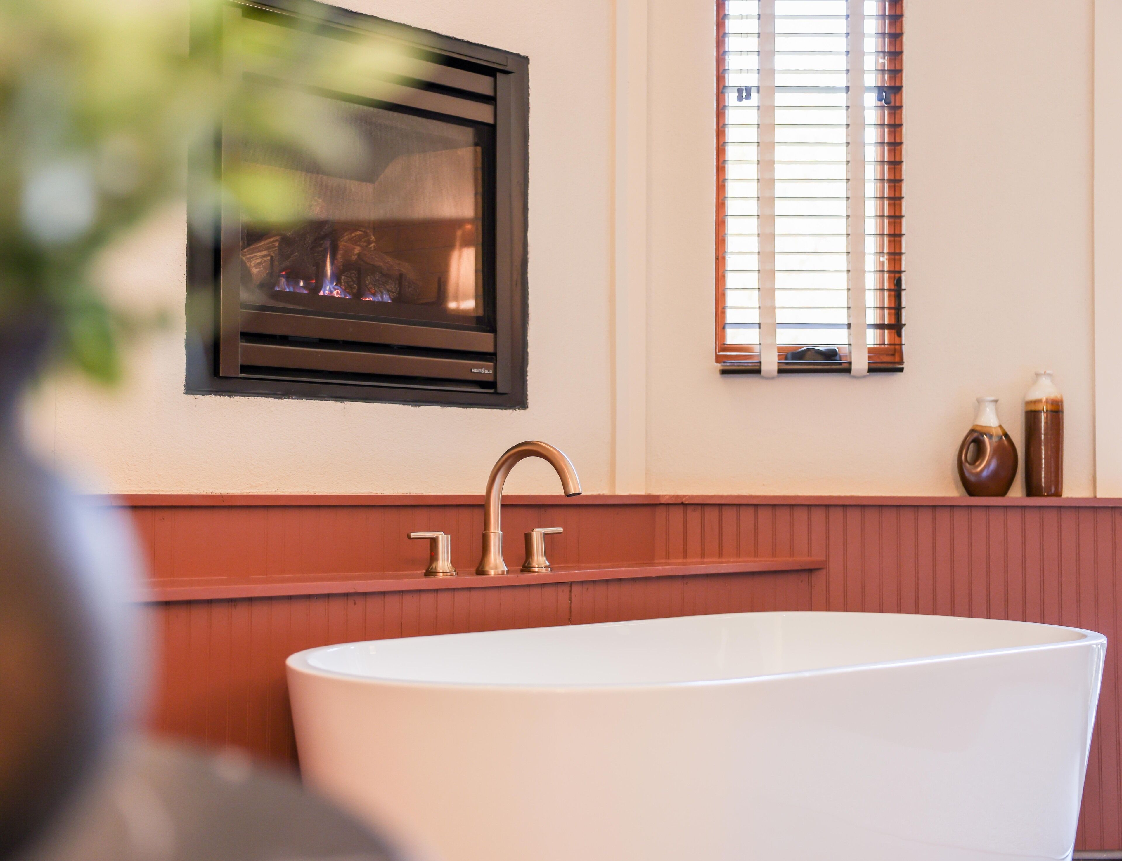 A modern bathroom with a freestanding bathtub, a wall-mounted TV, and a window with blinds.
