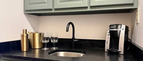 A kitchen with a black countertop, a Keurig coffee maker, two glasses, and a gold thermos.