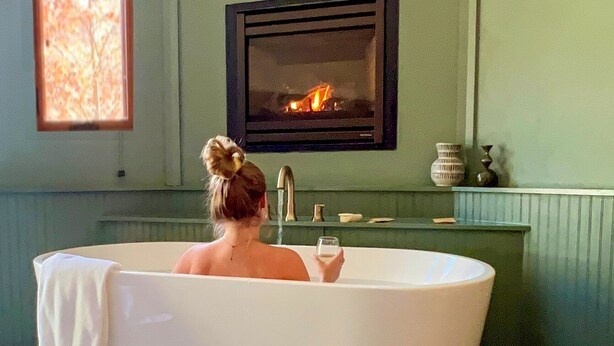 A woman sitting in a bathtub with a glass of milk, a fireplace, and a window with a view of trees.