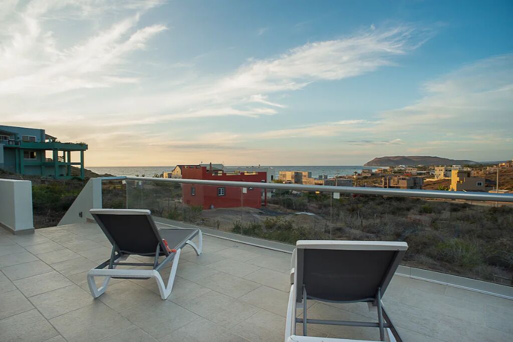 A balcony with two lounge chairs, a clear view of the sea, and buildings in the background.