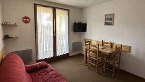 A room with a red sofa, a wooden dining table with chairs, a television on the wall, and a window with curtains.