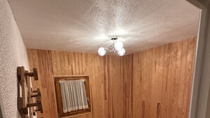 A small, cozy room with wooden walls, a single bed, a small window with curtains, and a ceiling light fixture.