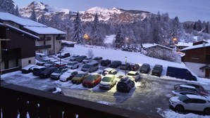 A snow-covered parking lot with cars and buildings, surrounded by snow-covered trees and mountains in the background.