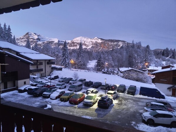 A snow-covered parking lot with cars and buildings, surrounded by snow-covered trees and mountains in the background.