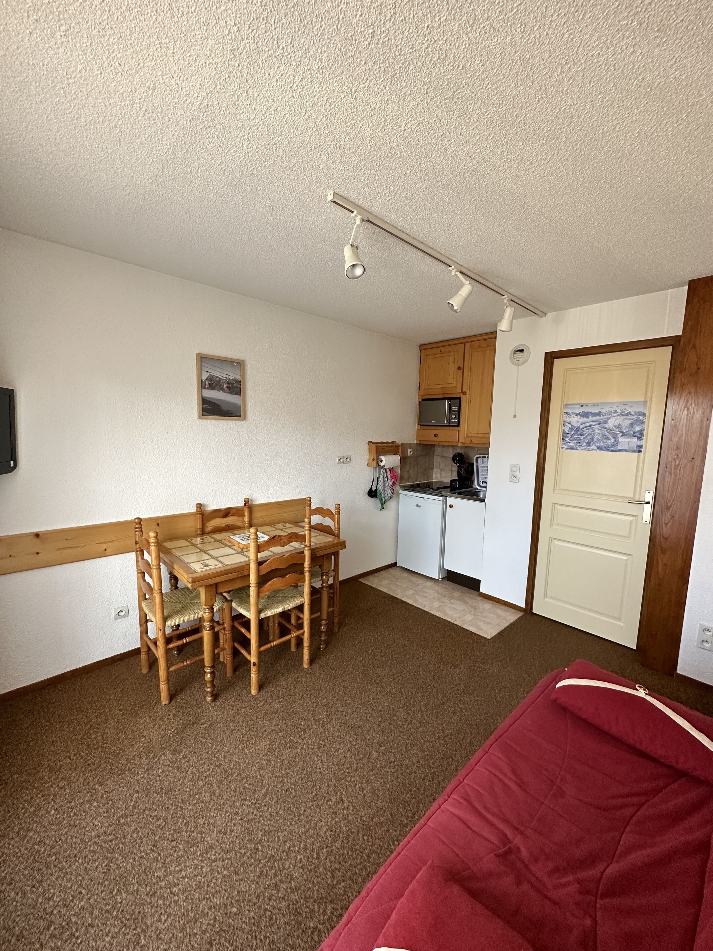 A compact room with a dining table and chairs, a kitchenette, a bed, and a door with a picture.