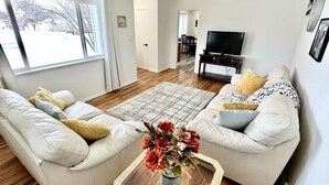 Una sala de estar con dos sofás blancos, una mesa de centro con un florero, un televisor sobre un mueble bajo y piso de madera.