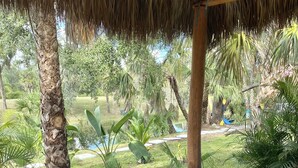 A covered outdoor seating area with two wicker lounge chairs, a thatched roof, and a view of a lush garden with various plants and a small pond. - Exotic waterfront Glamping experience  (Port Charlotte)
