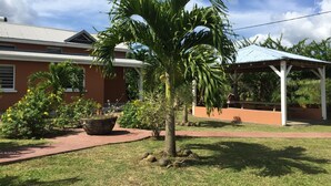 A building with a red exterior, a covered outdoor area, and a palm tree in the foreground. - Gîte Papaye · RESIDENCE LES KALBASSIERS (Lamentin)