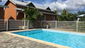 A swimming pool with stone tiles, surrounded by a white fence, and a building with a metal roof in the background. - Gîte Papaye · RESIDENCE LES KALBASSIERS (Lamentin)
