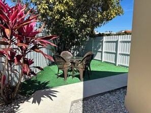 A patio area with a table and chairs, surrounded by a white fence and greenery.