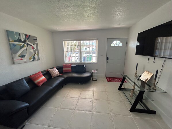 A living room with a black leather sofa, a glass coffee table, a painting on the wall, a door with a window, and a television mounted on the wall.