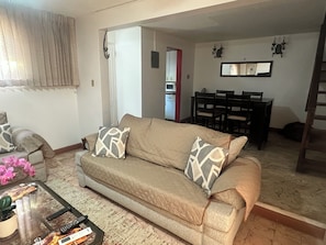 A living room with a beige sofa, a coffee table with a plant, and a dining area with a red cabinet and chairs.