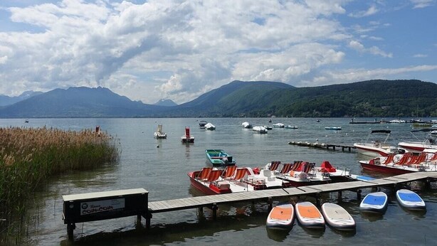 Un puerto deportivo con barcos amarrados, un muelle de madera y montañas al fondo.