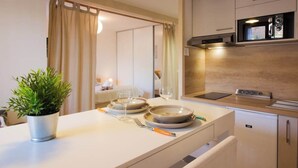 Cocina moderna con microondas, horno y un refrigerador pequeño. Hay un comedor con una mesa lista para dos, una planta en maceta y un espejo que refleja la habitación.