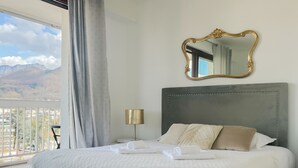A bedroom with a large bed, a nightstand, a lamp, and a mirror. There is a balcony with a view of mountains. - Mount Royal balcony with mountain view (Annecy)