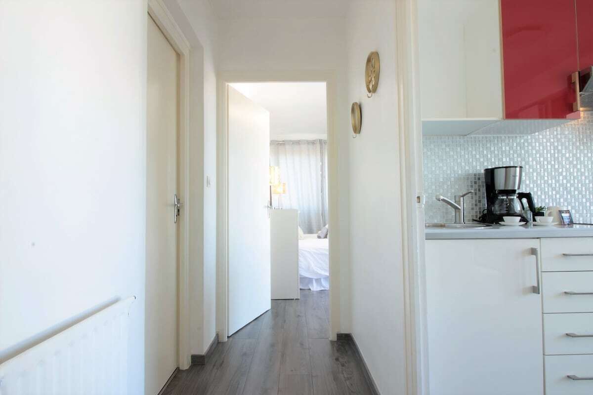 Un couloir étroit mène à une chambre à coucher qui contient un lit et une fenêtre avec des rideaux. On peut apercevoir une petite cuisine équipée d’une machine à café et d’un placard rouge.