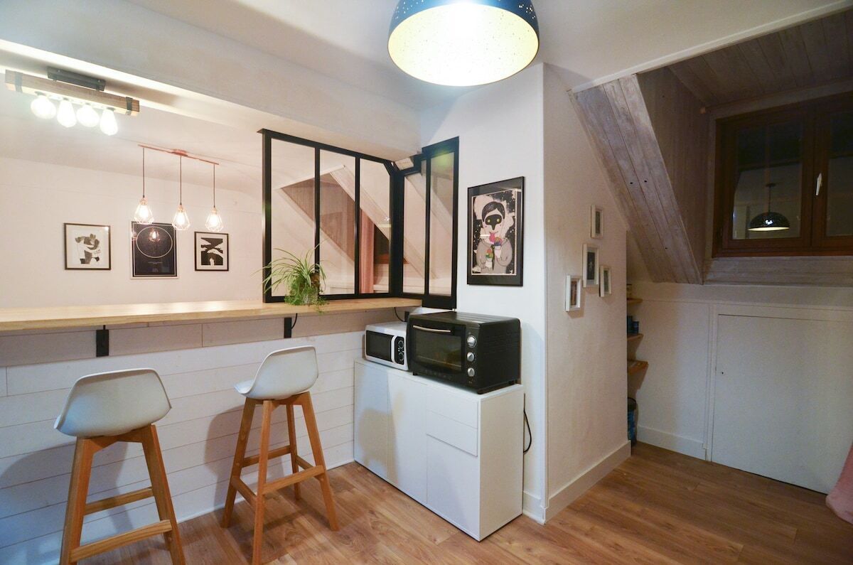 A compact kitchen with a microwave, two bar stools, a hanging light fixture, and framed artwork on the wall.