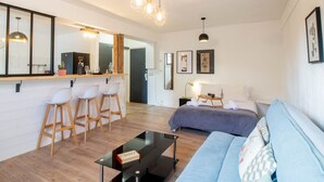 A modern living room with a sofa, a coffee table, and a bar area with stools. There is a bed with a blue blanket and a bedside table with a lamp. - Petit chez-vous pré carré rue Vaugelas (Annecy)