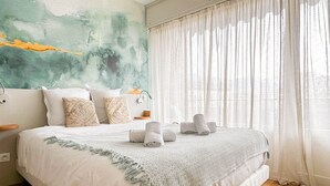 A neatly made bed with a white bedspread and a decorative throw, a bedside table with a lamp, and a large window with sheer curtains. - Central Park au centre ville, appareil raclette (Annecy)