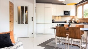 Une cuisine moderne avec des meubles blancs, un coin repas avec des chaises en bois et une grande fenêtre qui laisse entrer la lumière naturelle.