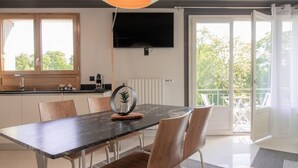 Une cuisine moderne avec un coin repas, un téléviseur fixé au-dessus du comptoir et un balcon offrant une vue sur les arbres.