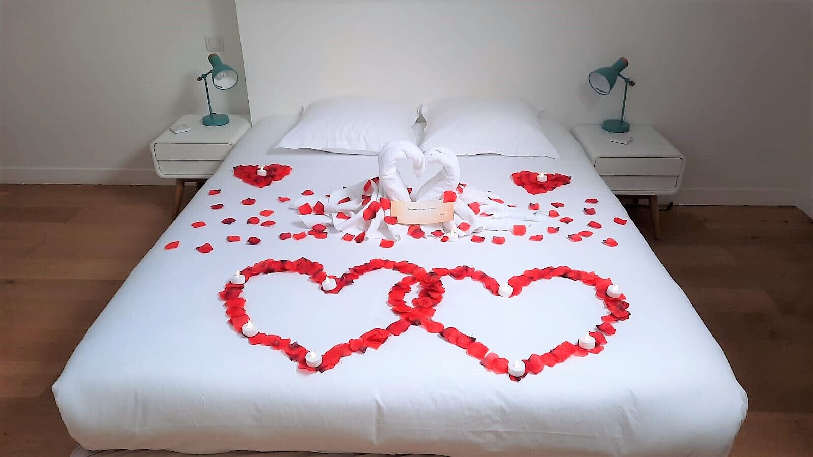 Een keurig opgemaakt bed met rozenblaadjes en twee zwanen, witte kussens en twee bedlampjes.