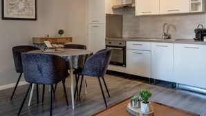 Eine moderne Küche mit weißen Schränken, einer Edelstahl-Dunstabzugshaube und einem eingebauten Backofen. Es gibt einen Essbereich mit gemusterten Stühlen und einem Tisch, und eine kleine Pflanze auf einem Holznachttisch.