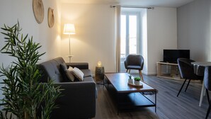 Ein modernes Wohnzimmer mit einer Couch, einem Couchtisch, Stühlen, einem Fernseher und einem großen Fenster mit Vorhängen.