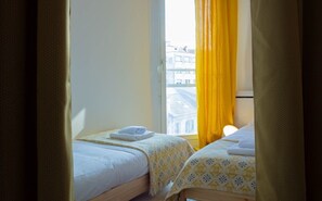 Quarto de hotel com duas camas, uma janela com cortinas amarelas e vista para prédios.