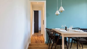 Un coin repas avec une table en bois dressée pour quatre personnes, deux lampes suspendues et une vue sur une autre pièce par une porte ouverte.