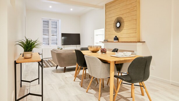 Un'area pranzo moderna con un tavolo in legno, quattro sedie, una pianta in vaso e una televisione sullo sfondo.