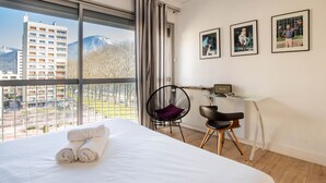 Ein Hotelzimmer mit einem großen Bett, einem Sessel, einem Schreibtisch und Blick auf Gebäude und Berge.