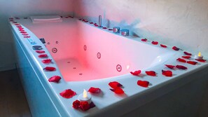 A rectangular bathtub with a pink interior, surrounded by red rose petals and candles. - Jacuzzi Bay (Annecy)