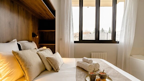 A hotel room with a bed, pillows, a tray with drinks and pastries, and a window with a view of buildings. - Jacuzzi Bay (Annecy)