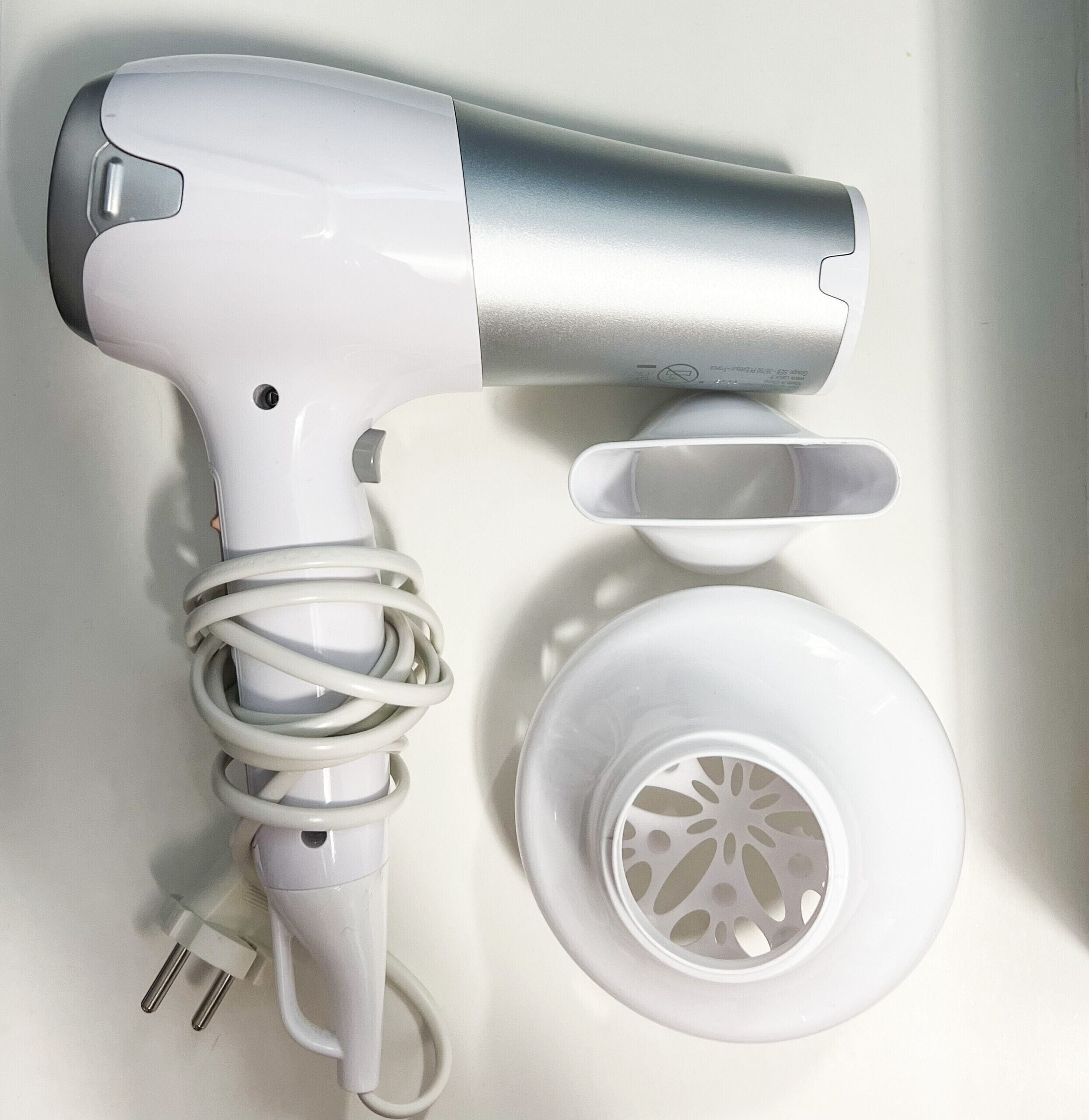 Un sèche-cheveux blanc avec un embout argenté, un cordon d’alimentation blanc et une prise électrique blanche.