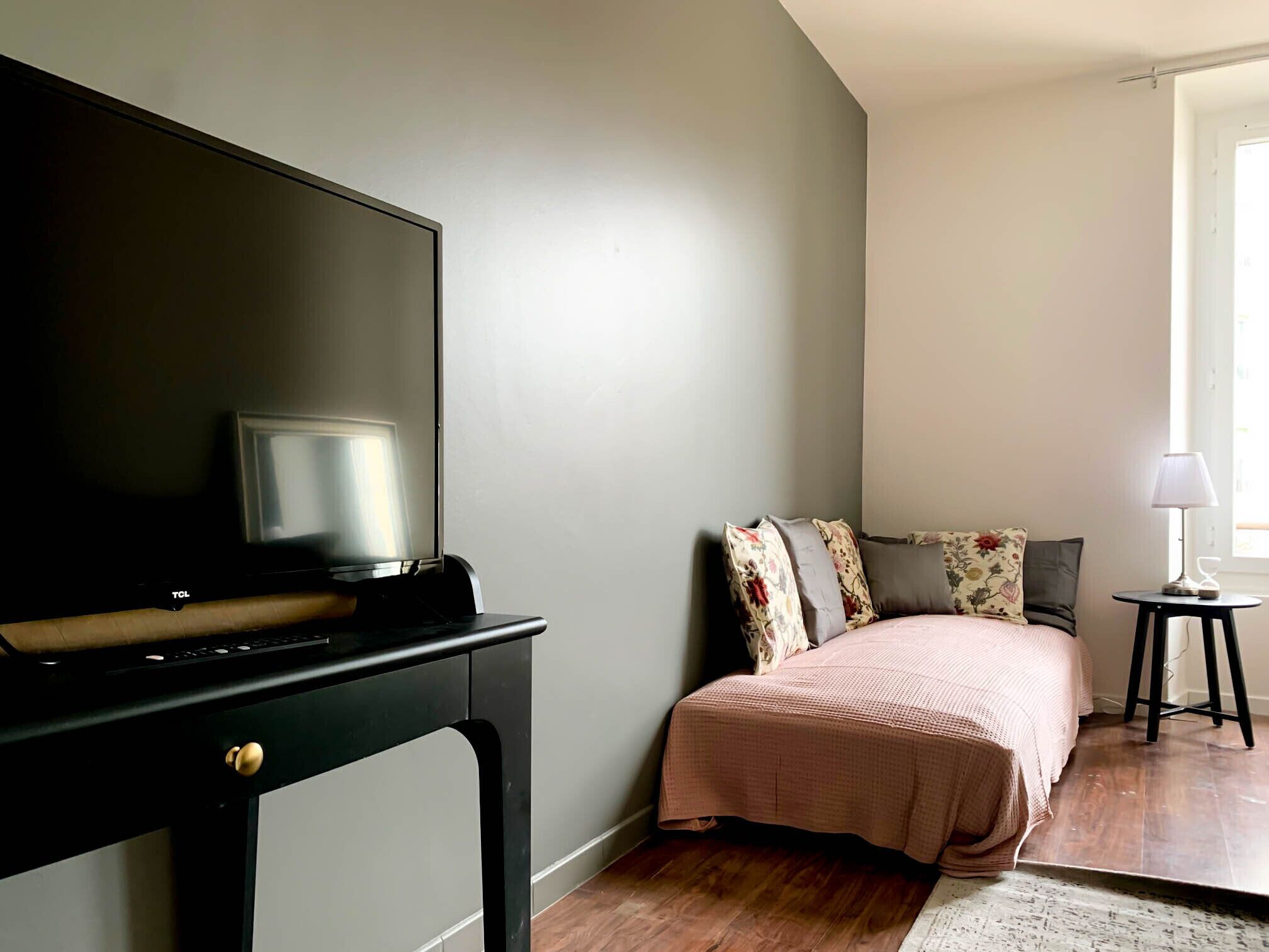 A bedroom with a bed, a TV on a stand, a small table with a lamp, and a window.