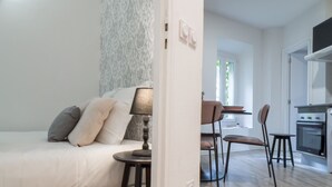 Ein modernes Schlafzimmer mit Bett, Nachttisch und Lampe. Es gibt eine Essecke mit Stühlen und einem Tisch. Der Raum verfügt über eine Küche mit Backofen und Schränken.