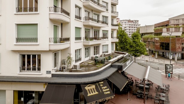 Ein mehrstöckiges Hotel mit Balkonen, Außenbestuhlung und einer Schaufensterfront.