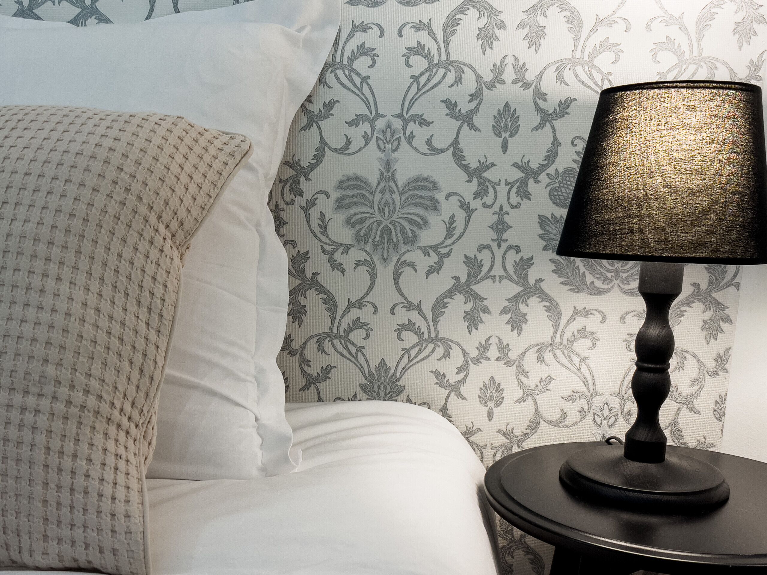 A bed with white linens and a textured blanket, a black lamp on a small table beside it, and a wall with a floral pattern.