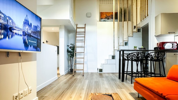 A modern living room with a flat-screen TV, wooden flooring, a staircase, and a kitchen area. - Atypique sous château vieille ville (Annecy)