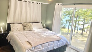 Un dormitorio con cama, mesitas de noche, ventilador de techo y vista a árboles a través de un ventanal grande.