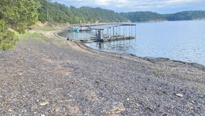 Una playa de guijarros con un muelle que se adentra en aguas tranquilas, rodeada de un bosque frondoso.
