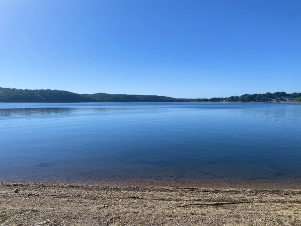 Ein stiller See mit blauem Himmel, umgeben von bewaldeter Landschaft und ein paar Gebäuden in der Ferne.