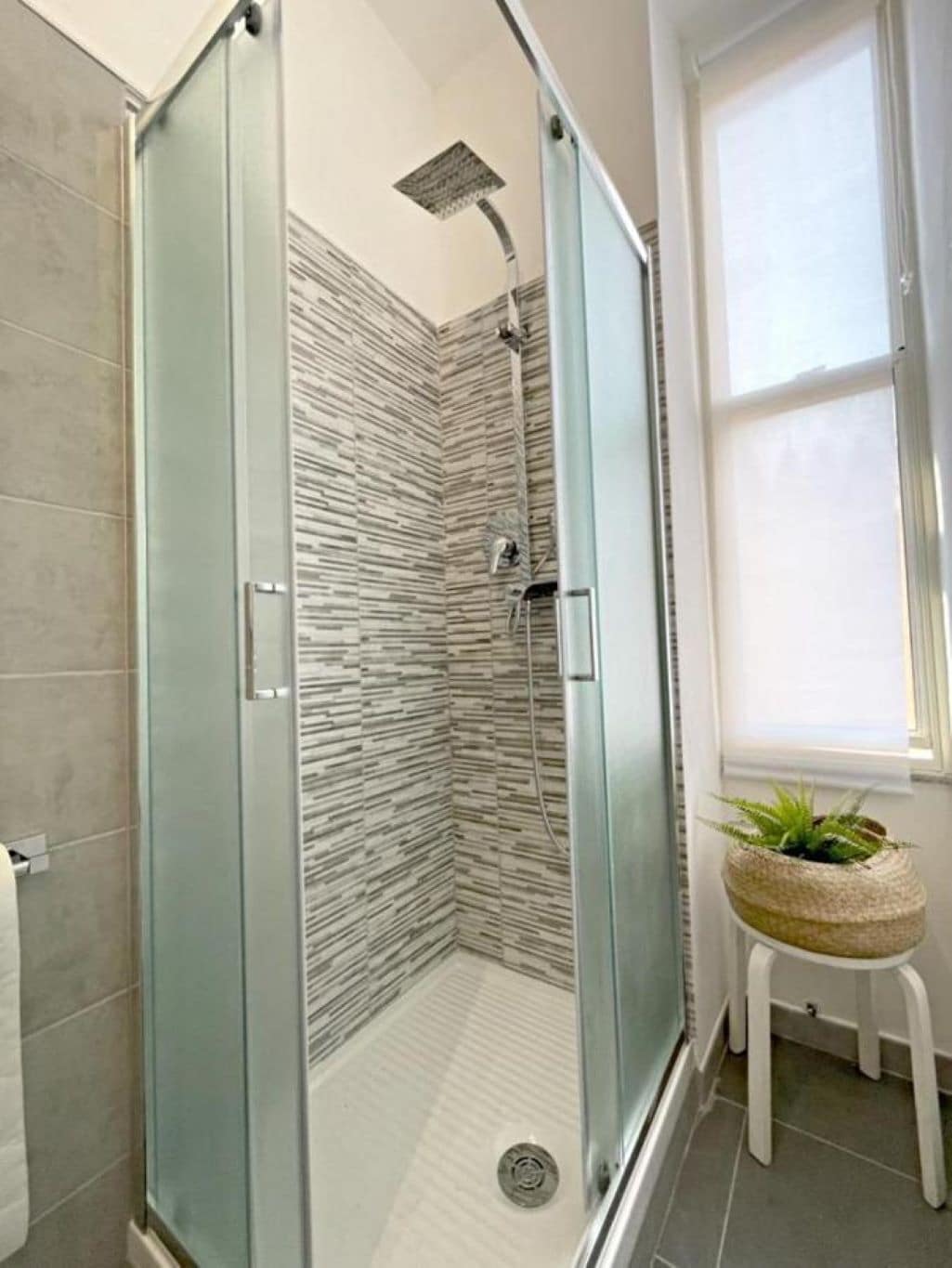 Une douche fermée par des parois en verre, avec un mur à texture, un petit tabouret agrémenté d’une plante et une fenêtre équipée de stores.