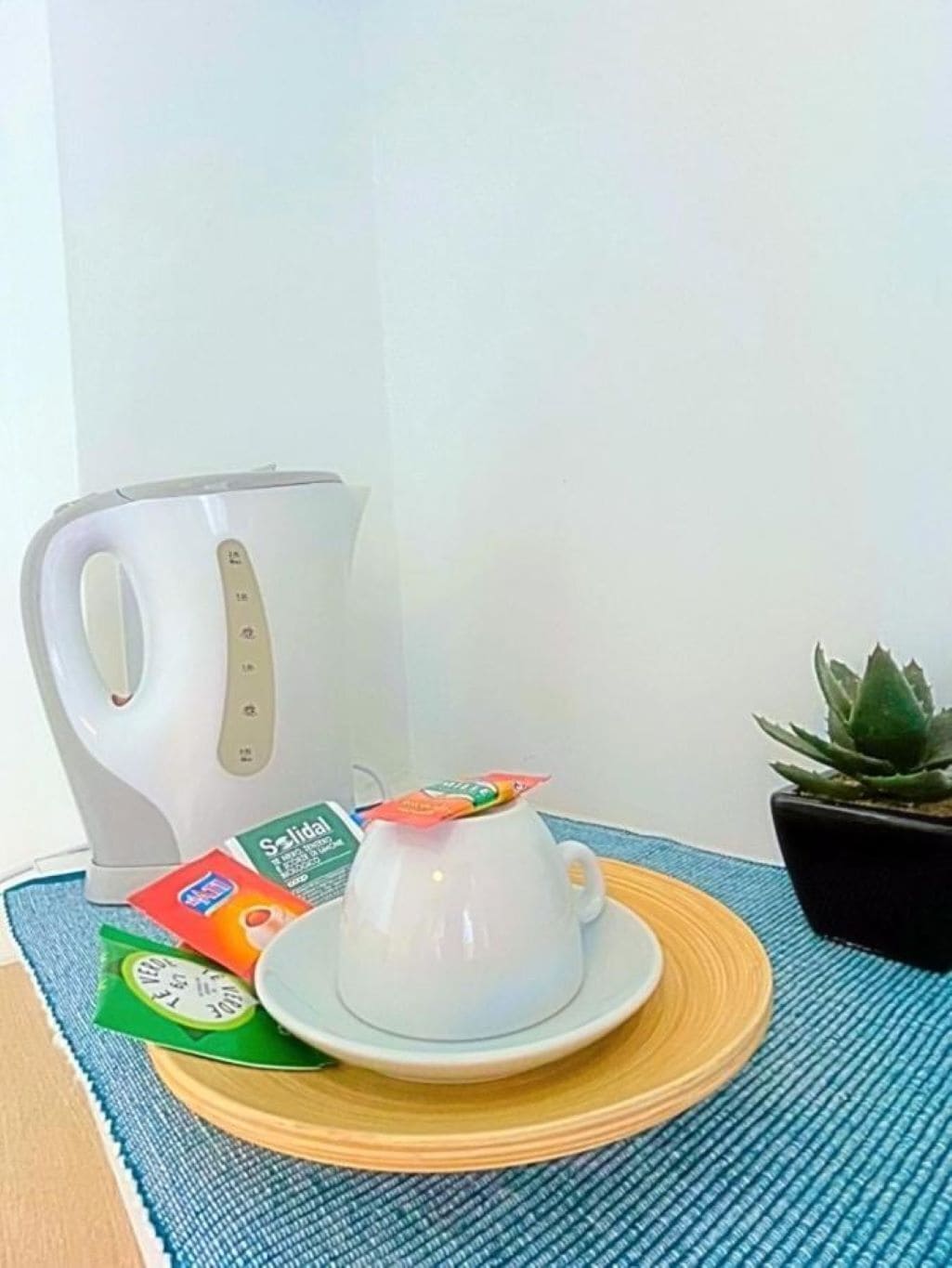 A white coffee maker, a white cup with a tea bag, and a small potted plant on a blue mat.
