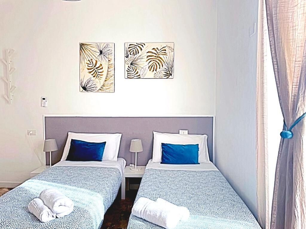 A hotel room with two single beds, each with a blue and white patterned bedspread, white pillows, and a blue accent pillow. There are two wall-mounted artworks depicting abstract leaf patterns. The room has a simple, clean design with a neutral color palette.