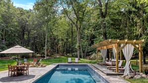 Una piscina rectangular con solárium de piedra, rodeada por una pérgola de madera con cortinas blancas, y un área de estar con sillas y una mesa.