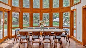 Un comedor amplio con una mesa y sillas de madera, ventanas grandes y un jarrón con flores.