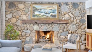 Una chimenea de piedra con un cuadro enmarcado encima, una zona de estar acogedora con una silla y un sofá, y una planta en maceta a la izquierda.