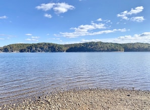 A calm lake with a pebbly shore and a forested hill in the background.